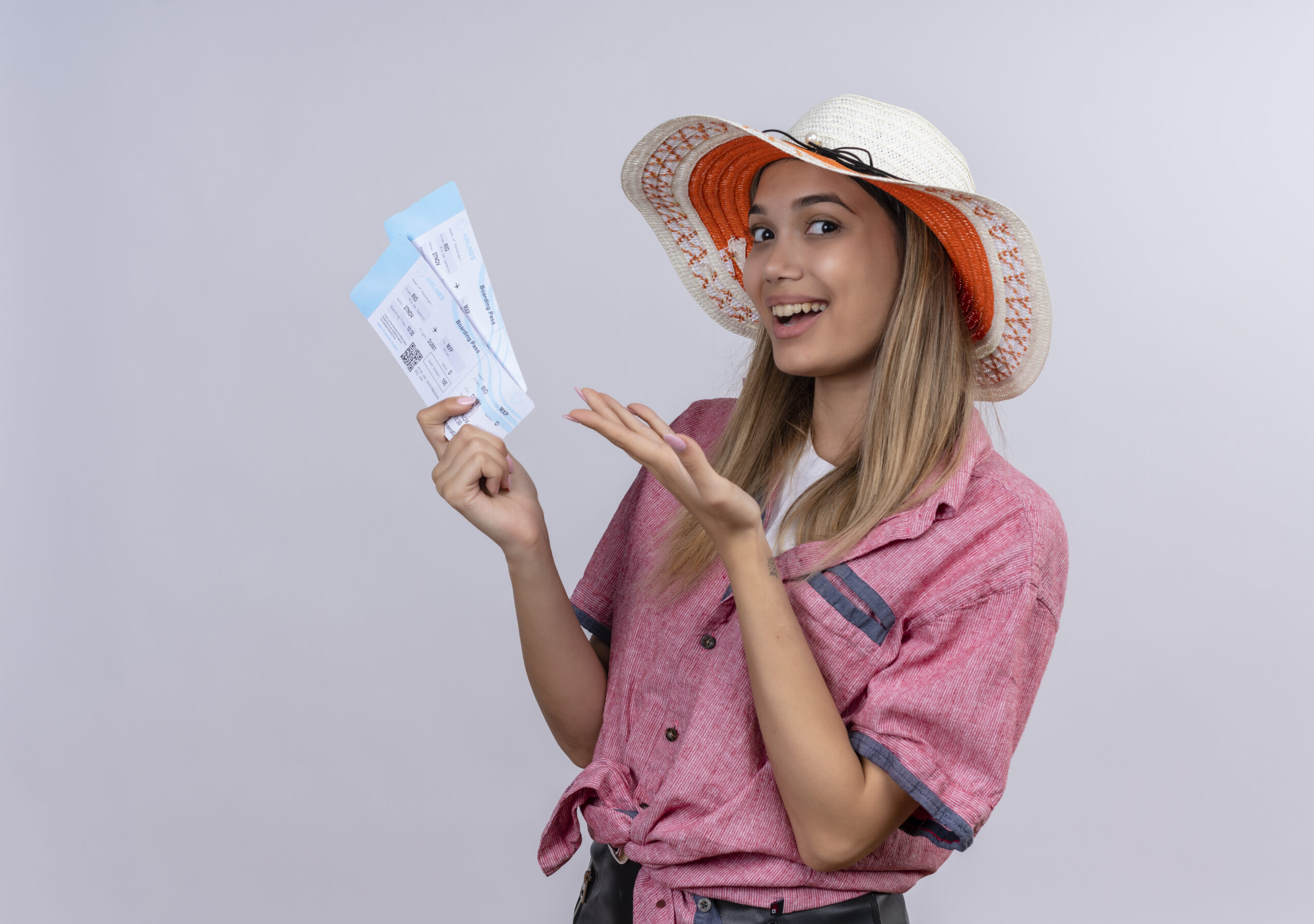 a happy young woman wearing red shirt and sun hat smiling and showing plane tickets on a white background